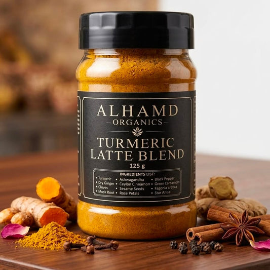 A jar of Alhamd Organics Turmeric Latte Blend sits on a wooden surface surrounded by ingredients like turmeric root, cinnamon, star anise, cloves, and loose turmeric powder. The background is a warm, blurred indoor setting.